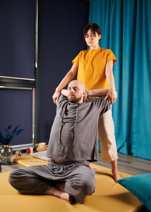 Masseuse making thai yoga massage. Woman therapist doing traditional massage treatment, stretching male patient's body. Bald client sitting on mat, enjoying hand massage in calm atmosphere.